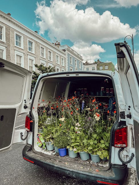 The open back of a white delivery van filled with a variety of freshly picked flowers, including red poppies, white daisies, and green foliage, arranged in small blue and grey pots. The flowers are vi