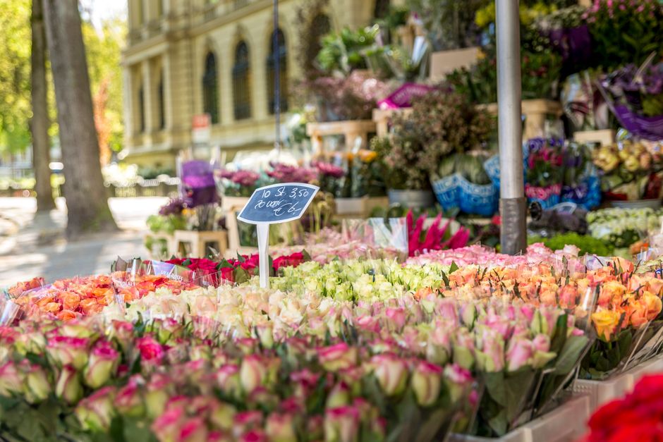 A vibrant outdoor flower stall displays a variety of fresh floral bouquets arranged in rows, featuring pastel pink, peach, white, and deep red roses. The flowers are wrapped in clear plastic and are p