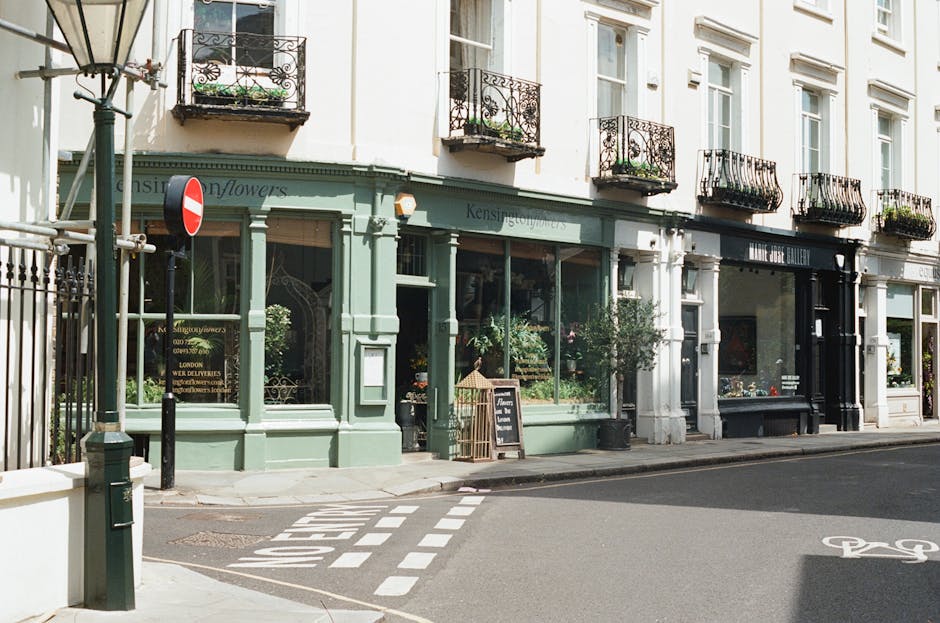 A view of a charming flower shop on England's Lane in Belsize Park, with a pastel green exterior and large glass windows showcasing an array of vibrant floral arrangements inside. The shop is surround