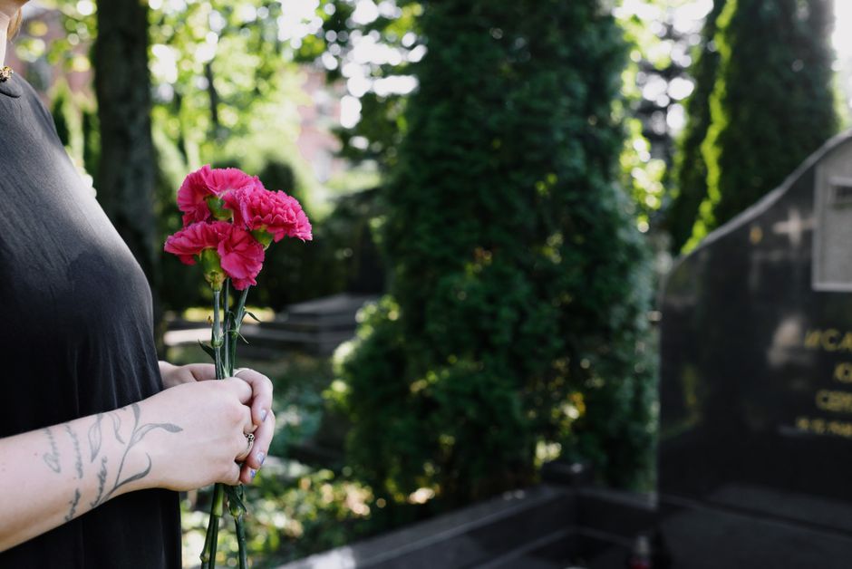 A person holding a small bouquet of bright pink carnations with ruffled petals and green stems, wrapped in clear plastic, standing outdoors near a grave with a polished black headstone. The person has