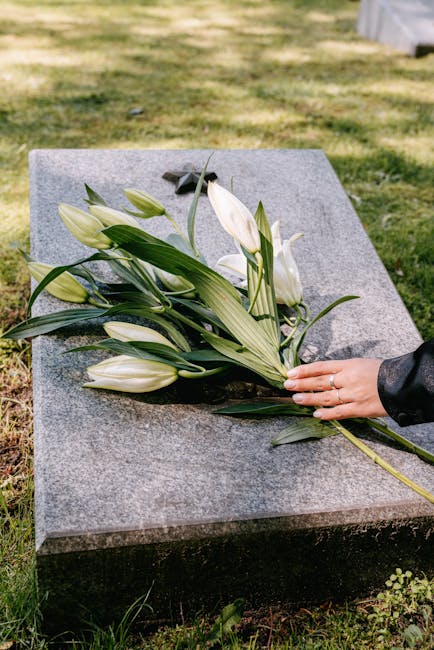 A person's hand, wearing a black leather jacket, gently places a bouquet of white lilies and green foliage onto a polished gray granite memorial stone. The arrangement features closed lily buds with s